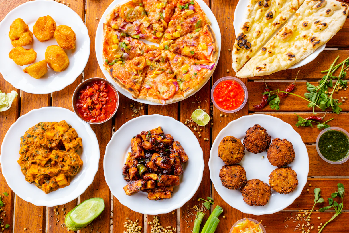 A variety of Indian dishes are arranged on a wooden table, including naan bread, pizza, fried snacks, and several curry dishes. Small bowls of colorful sauces accompany the plates, adding contrast to the vibrant display of food.