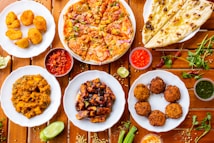 A variety of Indian dishes are arranged on a wooden table, including naan bread, pizza, fried snacks, and several curry dishes. Small bowls of colorful sauces accompany the plates, adding contrast to the vibrant display of food.