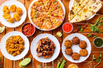 A variety of Indian dishes are arranged on a wooden table, including naan bread, pizza, fried snacks, and several curry dishes. Small bowls of colorful sauces accompany the plates, adding contrast to the vibrant display of food.