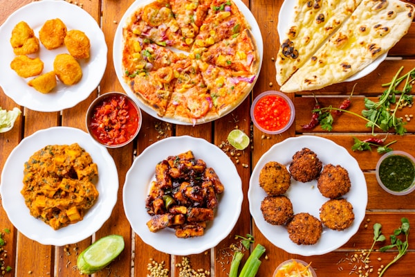 A variety of Indian dishes are arranged on a wooden table, including naan bread, pizza, fried snacks, and several curry dishes. Small bowls of colorful sauces accompany the plates, adding contrast to the vibrant display of food.