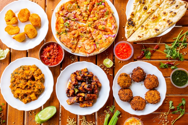 A variety of Indian dishes are arranged on a wooden table, including naan bread, pizza, fried snacks, and several curry dishes. Small bowls of colorful sauces accompany the plates, adding contrast to the vibrant display of food.