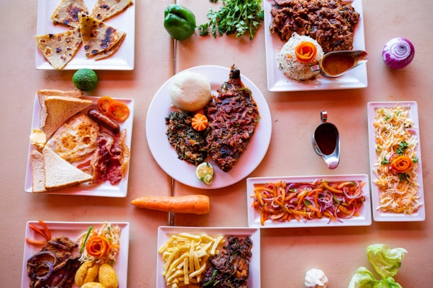 A diverse spread of dishes arranged on a tabletop, featuring plates with a mix of meats, vegetables, and sides. There are colorful garnishes and a variety of textures, indicating a meal likely associated with a special occasion or diverse menu tasting.