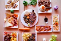 A diverse spread of dishes arranged on a tabletop, featuring plates with a mix of meats, vegetables, and sides. There are colorful garnishes and a variety of textures, indicating a meal likely associated with a special occasion or diverse menu tasting.