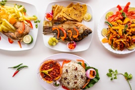 Four plates of diverse and colorful food are arranged on a white background. The first plate contains boiled whole fish paired with sliced red and yellow peppers and garnished with lime wedges. The second plate features chapati and a whole fried fish, accompanied by lime and pickled onions. The third plate has a variety of grilled vegetables with lime wedges. The fourth plate includes a dish of rice with raisins and beans, surrounded by garnishes like raw onion and a chili pepper. Fresh herbs and chili peppers are scattered across the table, enhancing the vibrant presentation.
