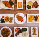 An assortment of various plated dishes arranged on a wooden table. The dishes include fish with spinach and staple food, a pastry, a meat dish with mashed potatoes and greens, a bowl of mixed vegetables with wraps, rice with garnishes, french fries with fried chicken, a vegetable and bean dish, a rib steak with spinach and rice, and samosas.