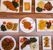 An assortment of various plated dishes arranged on a wooden table. The dishes include fish with spinach and staple food, a pastry, a meat dish with mashed potatoes and greens, a bowl of mixed vegetables with wraps, rice with garnishes, french fries with fried chicken, a vegetable and bean dish, a rib steak with spinach and rice, and samosas.