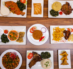An assortment of various plated dishes arranged on a wooden table. The dishes include fish with spinach and staple food, a pastry, a meat dish with mashed potatoes and greens, a bowl of mixed vegetables with wraps, rice with garnishes, french fries with fried chicken, a vegetable and bean dish, a rib steak with spinach and rice, and samosas.