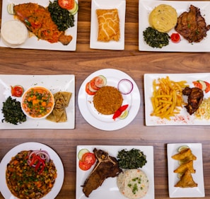 An assortment of various plated dishes arranged on a wooden table. The dishes include fish with spinach and staple food, a pastry, a meat dish with mashed potatoes and greens, a bowl of mixed vegetables with wraps, rice with garnishes, french fries with fried chicken, a vegetable and bean dish, a rib steak with spinach and rice, and samosas.