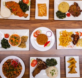 An assortment of various plated dishes arranged on a wooden table. The dishes include fish with spinach and staple food, a pastry, a meat dish with mashed potatoes and greens, a bowl of mixed vegetables with wraps, rice with garnishes, french fries with fried chicken, a vegetable and bean dish, a rib steak with spinach and rice, and samosas.