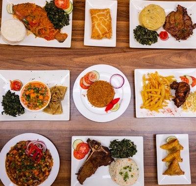An assortment of various plated dishes arranged on a wooden table. The dishes include fish with spinach and staple food, a pastry, a meat dish with mashed potatoes and greens, a bowl of mixed vegetables with wraps, rice with garnishes, french fries with fried chicken, a vegetable and bean dish, a rib steak with spinach and rice, and samosas.
