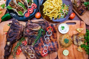 An arrangement of various foods is displayed on wooden cutting boards and plates. Grilled meat with rosemary and sliced onions, tacos with fresh cilantro and sliced tomatoes, a plate of fries with garnished vegetables, and a rice dish with a slice of lime are present. A bowl of white sauce accompanies the meal.