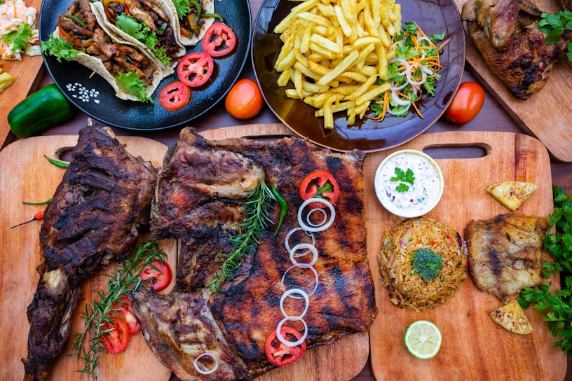 An arrangement of various foods is displayed on wooden cutting boards and plates. Grilled meat with rosemary and sliced onions, tacos with fresh cilantro and sliced tomatoes, a plate of fries with garnished vegetables, and a rice dish with a slice of lime are present. A bowl of white sauce accompanies the meal.