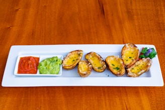 A rectangular white plate displays several potato skins topped with melted cheese and garnished with herbs. To the side, two square dishes contain a red dipping sauce and guacamole. The plate is on a wooden table, and there is a decorative garnish of fresh herbs with a purple flower on the right side.