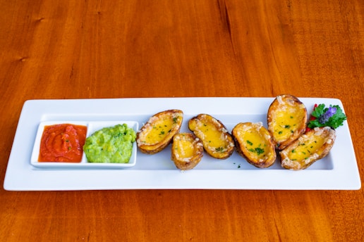 A rectangular white plate displays several potato skins topped with melted cheese and garnished with herbs. To the side, two square dishes contain a red dipping sauce and guacamole. The plate is on a wooden table, and there is a decorative garnish of fresh herbs with a purple flower on the right side.