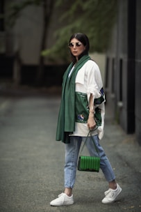 A stylishly dressed person wearing sunglasses, a green scarf, and a white top with unique embroidery on the sleeves. Holding a green handbag, the person is also wearing light blue jeans and white sneakers, standing on a paved walkway with a blurred, natural background.