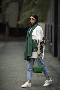 A stylishly dressed person wearing sunglasses, a green scarf, and a white top with unique embroidery on the sleeves. Holding a green handbag, the person is also wearing light blue jeans and white sneakers, standing on a paved walkway with a blurred, natural background.