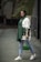 A stylishly dressed person wearing sunglasses, a green scarf, and a white top with unique embroidery on the sleeves. Holding a green handbag, the person is also wearing light blue jeans and white sneakers, standing on a paved walkway with a blurred, natural background.