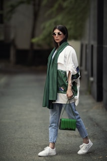 A stylishly dressed person wearing sunglasses, a green scarf, and a white top with unique embroidery on the sleeves. Holding a green handbag, the person is also wearing light blue jeans and white sneakers, standing on a paved walkway with a blurred, natural background.