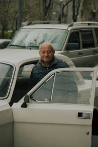 An elderly person stands beside an opened car door, looking directly into the camera with a gentle smile. Behind the person, there is another car, and in the background, trees and other elements of an urban environment can be seen.