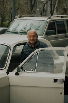 An elderly person stands beside an opened car door, looking directly into the camera with a gentle smile. Behind the person, there is another car, and in the background, trees and other elements of an urban environment can be seen.