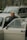 An elderly person stands beside an opened car door, looking directly into the camera with a gentle smile. Behind the person, there is another car, and in the background, trees and other elements of an urban environment can be seen.