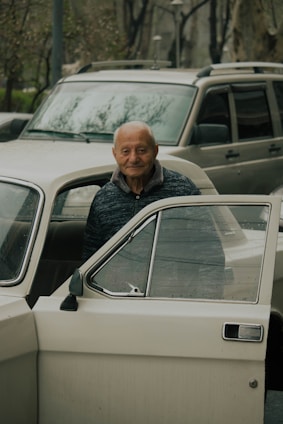 An elderly person stands beside an opened car door, looking directly into the camera with a gentle smile. Behind the person, there is another car, and in the background, trees and other elements of an urban environment can be seen.