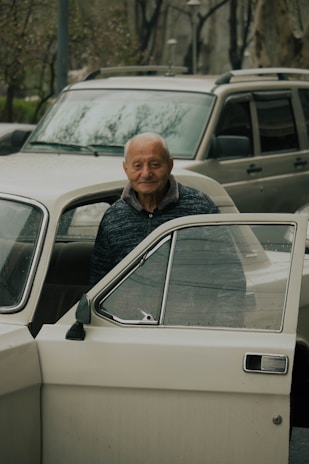 An elderly person stands beside an opened car door, looking directly into the camera with a gentle smile. Behind the person, there is another car, and in the background, trees and other elements of an urban environment can be seen.