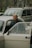 An elderly person stands beside an opened car door, looking directly into the camera with a gentle smile. Behind the person, there is another car, and in the background, trees and other elements of an urban environment can be seen.