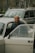 An elderly person stands beside an opened car door, looking directly into the camera with a gentle smile. Behind the person, there is another car, and in the background, trees and other elements of an urban environment can be seen.