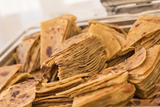 A heap of sliced, golden-brown flatbread stacked neatly, showcasing layers and slightly charred surfaces. The bread appears crispy and is arranged in a metallic tray, suggesting a setting such as a buffet or a dining setup.