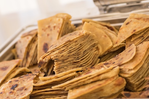 A heap of sliced, golden-brown flatbread stacked neatly, showcasing layers and slightly charred surfaces. The bread appears crispy and is arranged in a metallic tray, suggesting a setting such as a buffet or a dining setup.