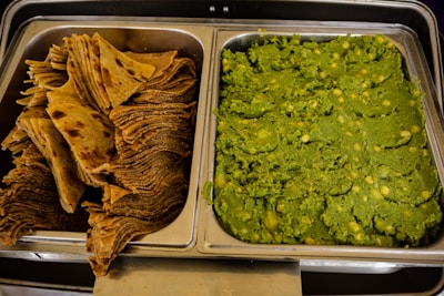 A serving tray containing two dishes side by side: on the left are neatly stacked brown, crispy flatbreads, and on the right is a tray filled with a vibrant green mashed mixture, likely containing peas or other legumes.