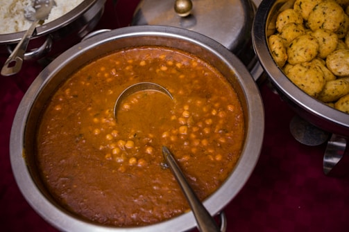 A serving dish filled with chickpea curry, surrounded by other dishes including rice and round, yellow flatbreads with herbs. The chickpea dish has a metal ladle in it and is presented on a dark red tablecloth.
