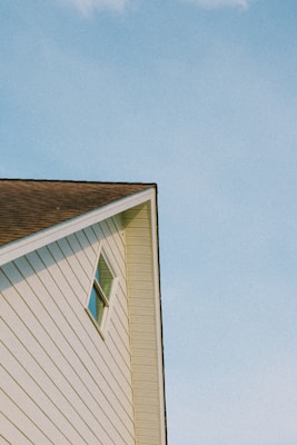 A white triangular gable of a house with a small rectangular window is set against a clear blue sky. The siding is vertical with uniform lines, and the roof is steeply pitched with brown shingles.