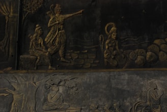 The image displays a detailed stone relief depicting Hindu mythological figures and scenes. A central figure is pointing forward, accompanied by another seated figure and a monkey-like figure carrying stones. Below, there is a scene with a figure seated under a tree in meditation.