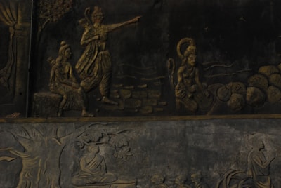 The image displays a detailed stone relief depicting Hindu mythological figures and scenes. A central figure is pointing forward, accompanied by another seated figure and a monkey-like figure carrying stones. Below, there is a scene with a figure seated under a tree in meditation.
