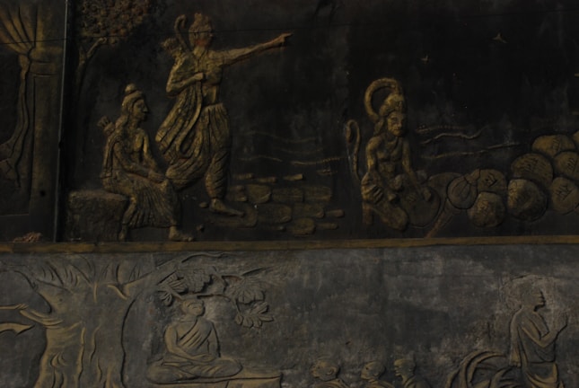 The image displays a detailed stone relief depicting Hindu mythological figures and scenes. A central figure is pointing forward, accompanied by another seated figure and a monkey-like figure carrying stones. Below, there is a scene with a figure seated under a tree in meditation.