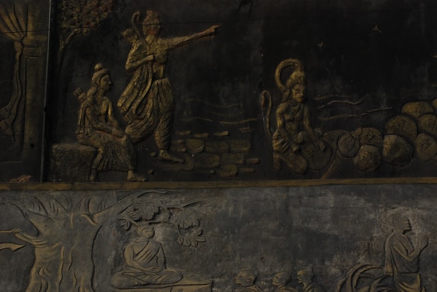 The image displays a detailed stone relief depicting Hindu mythological figures and scenes. A central figure is pointing forward, accompanied by another seated figure and a monkey-like figure carrying stones. Below, there is a scene with a figure seated under a tree in meditation.