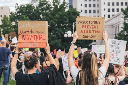 A group of people are gathered outdoors in a protest. Several individuals are holding up signs with messages, such as 'Girls just want to have fundamental rights' and 'Vasectomies prevent abortion.' In the background, more protest signs and a mix of trees and a building are visible.