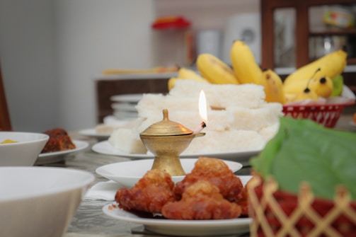 A dining table adorned with various food items including bananas and fried pastries. A lit brass oil lamp is centrally placed. The background includes plates stacked together and other blurred utensils.