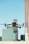 A drone is flying in the foreground with its wings extended, set against a blurred urban backdrop featuring tall buildings under a clear blue sky.