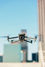 A drone is flying in the foreground with its wings extended, set against a blurred urban backdrop featuring tall buildings under a clear blue sky.