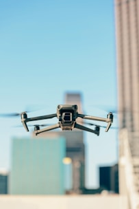 A drone is flying in the foreground with its wings extended, set against a blurred urban backdrop featuring tall buildings under a clear blue sky.