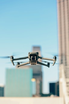 A drone is flying in the foreground with its wings extended, set against a blurred urban backdrop featuring tall buildings under a clear blue sky.