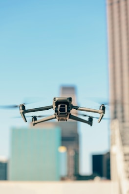 A drone is flying in the foreground with its wings extended, set against a blurred urban backdrop featuring tall buildings under a clear blue sky.