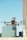 A drone is flying in the foreground with its wings extended, set against a blurred urban backdrop featuring tall buildings under a clear blue sky.
