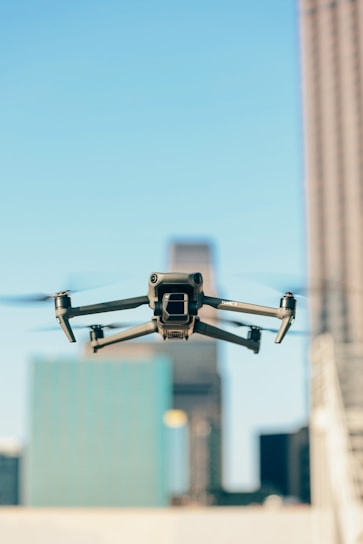 A drone is flying in the foreground with its wings extended, set against a blurred urban backdrop featuring tall buildings under a clear blue sky.