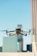 A drone is flying in the foreground with its wings extended, set against a blurred urban backdrop featuring tall buildings under a clear blue sky.