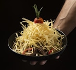A black bowl is filled with a stack of thin, crispy fried potato sticks, topped with a cherry tomato and a sprig of rosemary. The bowl is held by a person's hand against a dark background.