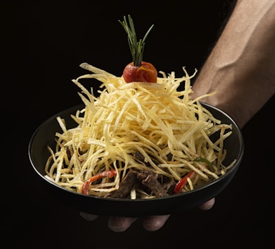 A black bowl is filled with a stack of thin, crispy fried potato sticks, topped with a cherry tomato and a sprig of rosemary. The bowl is held by a person's hand against a dark background.